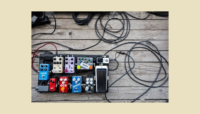 A pedalboard with a grid layout, showing multiple rows of guitar pedals including drive, modulation, and delay effects, with cables coiled around it on a wooden stage floor.
