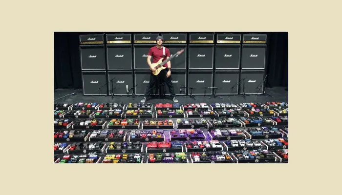 Guitarist standing in front of a massive wall of Marshall amps and an oversized pedalboard filled with hundreds of effects pedals—an exaggerated example of going too big for most players’ needs.