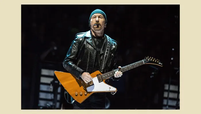 The Edge from U2 performing live with orange Gibson Explorer guitar, wearing black leather jacket and his signature beanie